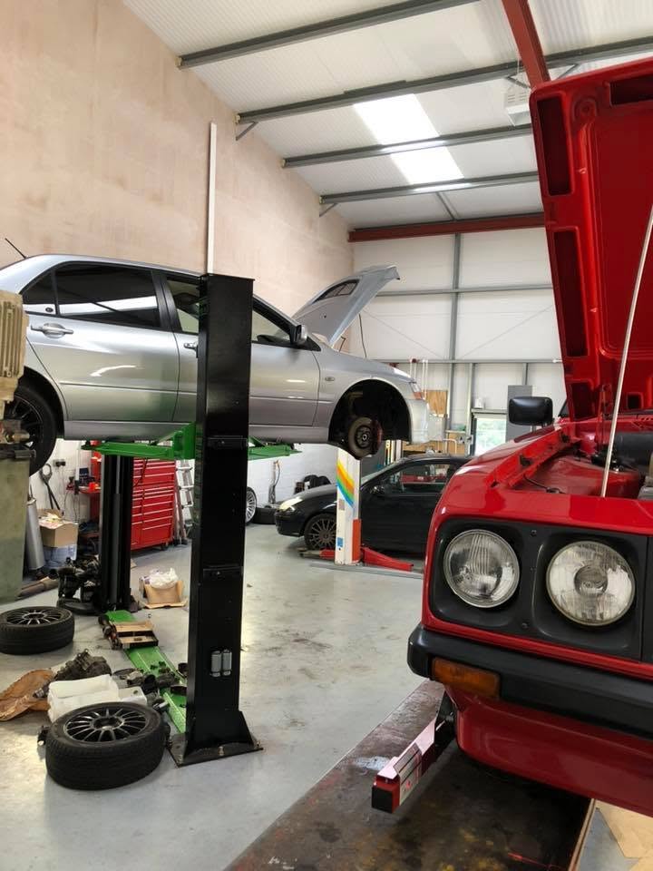 A car raised on a two-post lift for repairs, next to a classic with its bonnet up
